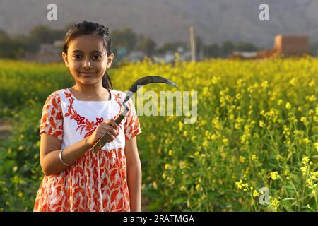 Ein indisches Bauernmädchen, das auf dem Senffeld steht und ein Kleid trägt. Ein Mädchen genießt die blühende Ernte mit Sichel in der Hand. Stockfoto