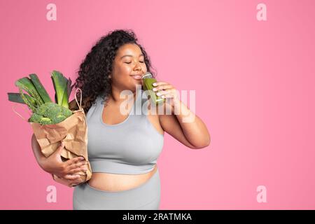 Fröhliche schwarze, übergroße Dame in Sportbekleidung, trinkt Smoothie, hält eine Tasche mit grünem Gemüse, rosa Hintergrund, Kopierraum Stockfoto