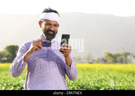 Ein glücklicher indischer Bauer auf dem Land, der auf dem Senffeld steht und Kurta-Pyjama trägt. Online-Zahlung per Smartphone mit Kreditkarte. Stockfoto