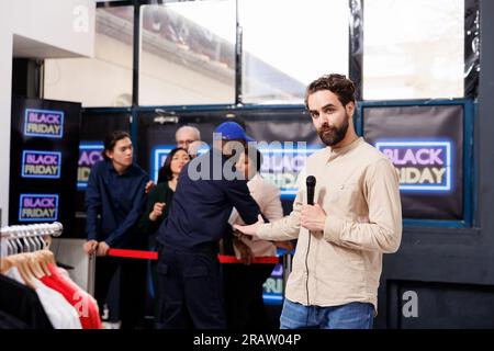 Journalistin mit Mikrophon-Geste, während sie Black Friday Reportage in der Modeboutique machte. TV-Reporter, der über Mikrofon spricht, während er gegen die Menge von Käufern steht, die gerne einkaufen möchten Stockfoto