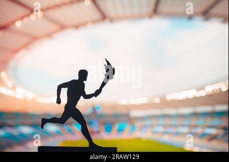 Den Geist entzünden: Silhouette eines männlichen Athleten, der das Fackelrelais trägt. Modernes Leichtathletik-Stadion als markante Kulisse. Das Essen einfangen Stockfoto