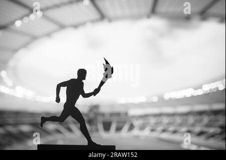 Passing the Flame: Silhouette des männlichen Athleten in Torch Relay, vor einem modernen Leichtathletik-Stadion. Ein beeindruckendes Bild für das Sommerevent 2024 Stockfoto