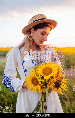 Porträt einer jungen Frau, die am blühenden Sonnenblumenfeld bei Sonnenuntergang vorbeigeht, mit Blumenstrauß und Landschaft. Model trägt ethnische Kleidung und ha Stockfoto