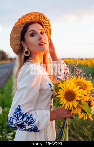 Porträt einer jungen Frau, die am blühenden Sonnenblumenfeld bei Sonnenuntergang vorbeigeht, mit Blumenstrauß und Natur. Model trägt ethnisch gestickte Kleidung Stockfoto