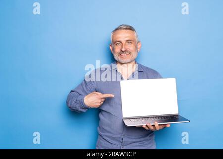 Glücklicher Kaukasier mittleren Alters zeigt Finger auf Laptop mit leerem Bildschirm, empfiehlt Computer, isoliert auf blauem Hintergrund. Covid-19 Quarantäne mit m Stockfoto