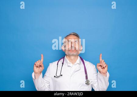 Mittelalter schöner Arzt Mann trägt Mantel und Stethoskop über isolierten blauen Hintergrund lächelnd mit einer Idee oder Frage zeigt Finger nach oben mit Hap Stockfoto