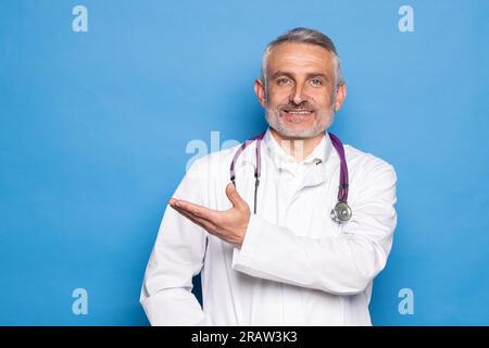 Hübscher Arzt mit Mantel und Stethoskop auf isoliertem blauem Hintergrund Stockfoto