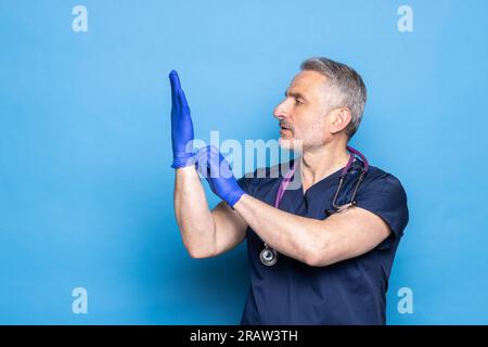Reifer männlicher Arzt in medizinischer Uniform Anlegen steriler blauer Schutzhandschuhe, während er vor blauem Hintergrund steht. Stockfoto