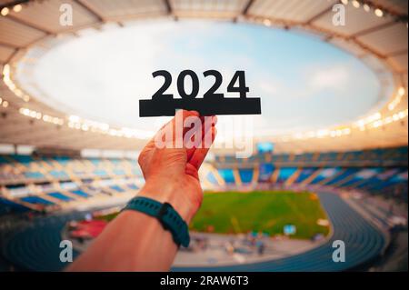 Silhouette von „2024“ in der Hand von Athleten. Das Zeichen signalisiert den Beginn des Sportjahres und führt zu den Sommerspielen in Paris. Modernes Sportstadion hinten Stockfoto