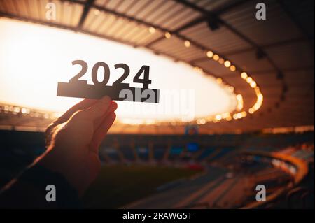 Silhouette von „2024“ in der Hand von Athleten. Das Zeichen signalisiert den Beginn des Sportjahres und führt zu den Sommerspielen in Paris. Modernes Sportstadion hinten Stockfoto