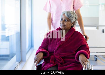 Vielfältige Ärztin und ältere Patientin im Rollstuhl, die durch das Fenster im Krankenhaus blickt. Krankenhaus, Medizin und Gesundheitswesen, unalter Stockfoto