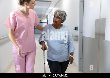 Vielfältige Ärztin und ältere Patientin mit Krücke, die im Flur im Krankenhaus läuft. Krankenhaus-, Medizin- und Gesundheitsdienstleistungen, unverändert. Stockfoto