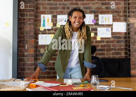 Porträt eines birassischen männlichen Modedesigners mit Dreadlocks auf dem Schreibtisch, die im Studio lächeln. Geschäft, Arbeit, Selbstvertrauen, Mode und Design, unverfälscht Stockfoto