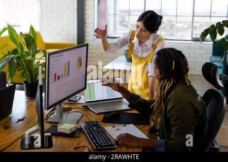 Verschiedene Gelegenheitsarbeitskollegen, die Computer und Laptops nutzen, um am Schreibtisch im Büro über ihre Arbeit zu sprechen. Teamwork, Arbeit, Arbeit, Kommunikation und Freizeit Stockfoto