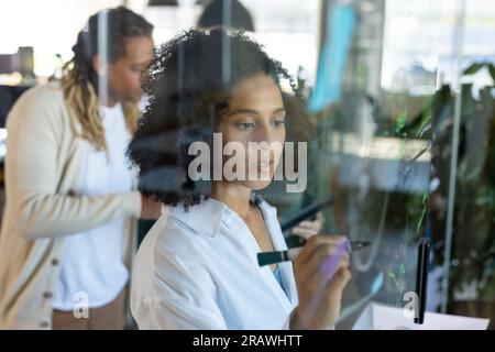 Fokussierte, birassische, zwanglose Geschäftsfrau, die Brainstorming mit Kollegen führt und Notizen an Glaswänden macht. Teamwork, Arbeit, Business, Brainstorming, Meeting und cas Stockfoto