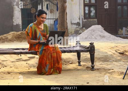 Frau, die im ländlichen Raum sitzt und Saree trägt, Sari, ein traditionelles Kleid für Frauen in Nordindien, mit einem Laptop in der Hand. Stockfoto