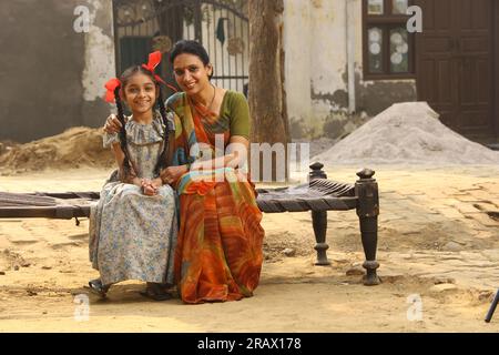 Glückliche indische Landfamilie im Dorf. Mutter und Töchter sitzen zusammen und lächeln auf einem Kinderbett vor ihrem Haus im Vorgarten. Mutter in wunderschöner Saree Stockfoto