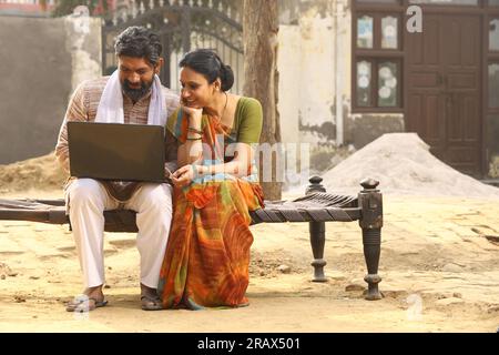 Glückliche indische Landfamilie im Dorf. Ländliches Paar sitzt zusammen und lächelt auf dem Kinderbett vor dem Haus und benutzt einen Laptop für Online-Zahlungen. Stockfoto