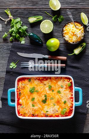 Vegetarische Enchilada-Kasserolle mit Tortillas, Sauce, Pinto-Bohnen, Mais, Zucchini und mexikanischem Käse in Backform auf dunklem Holztisch, Stockfoto