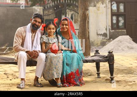 Glückliche ländliche indische Familie, die zusammensitzt, hält die Tochter ein Sparschwein in der Hand, steckt eine Münze hinein und zeigt das Konzept der Geldeinsparung Stockfoto