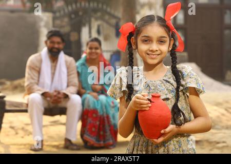 Glückliche ländliche indische Familie, die zusammensitzt, hält die Tochter ein Sparschwein in der Hand, steckt eine Münze hinein und zeigt das Konzept der Geldeinsparung Stockfoto