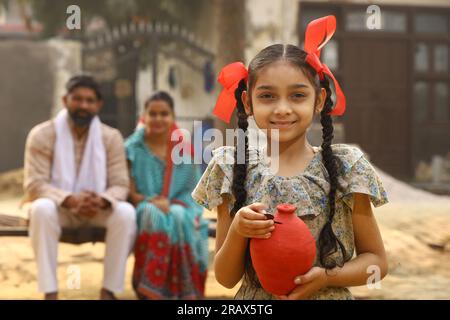 Glückliche ländliche indische Familie, die zusammensitzt, hält die Tochter ein Sparschwein in der Hand, steckt eine Münze hinein und zeigt das Konzept der Geldeinsparung Stockfoto