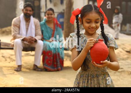 Glückliche ländliche indische Familie, die zusammensitzt, hält die Tochter ein Sparschwein in der Hand, steckt eine Münze hinein und zeigt das Konzept der Geldeinsparung Stockfoto