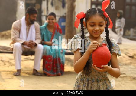 Glückliche ländliche indische Familie, die zusammensitzt, hält die Tochter ein Sparschwein in der Hand, steckt eine Münze hinein und zeigt das Konzept der Geldeinsparung Stockfoto