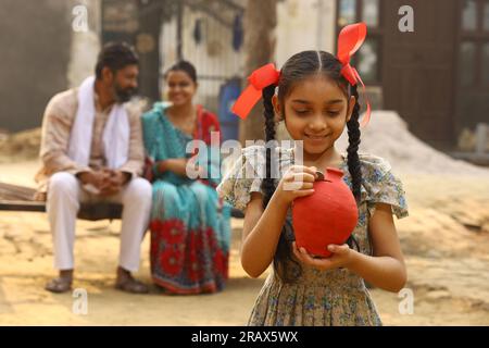 Glückliche ländliche indische Familie, die zusammensitzt, hält die Tochter ein Sparschwein in der Hand, steckt eine Münze hinein und zeigt das Konzept der Geldeinsparung Stockfoto