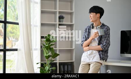 Ein glücklicher und gutaussehender junger asiatischer Freiberufler in einem Flanellhemd steht in seinem Büro mit einer Kaffeetasse in der Hand und blickt Tag und Nacht aus dem Fenster Stockfoto