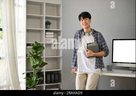 Ein hübscher und glücklicher junger Asiate in einem Flanellhemd steht in seinem modernen Büro mit Büchern in der Hand. Stockfoto