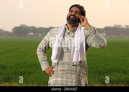 Glücklicher Bauer auf dem Land Indiens, der sein Mobiltelefon benutzt, telefoniert mit seinem Smartphone, während er im Morgengrauen auf einem landwirtschaftlichen Feld steht Stockfoto