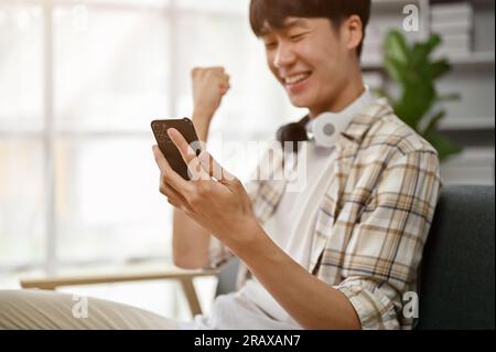 Nahaufnahme eines überfreulichen und glücklichen jungen Asiaten, der auf seinen Smartphone-Bildschirm schaut und seine guten Nachrichten feiert, während er auf dem Sofa sitzt Stockfoto