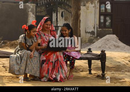 Glückliche indische Landfamilie im Dorf. Mutter und Töchter sitzen zusammen und lächeln auf einem Kinderbett vor ihrem Haus im Vorgarten. Mutter in wunderschöner Saree Stockfoto