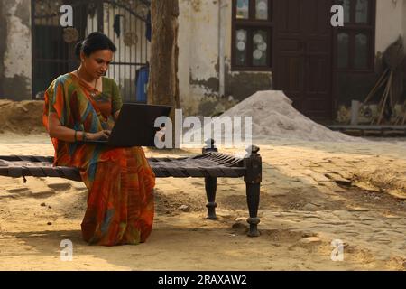 Glückliche indische Landfrau im Dorf. Sie sitzt auf dem Kinderbett und lächelt draußen in einem wunderschönen Saree. Indische Kultur, traditionelle Kleidung, Surfen auf einem Laptop Stockfoto