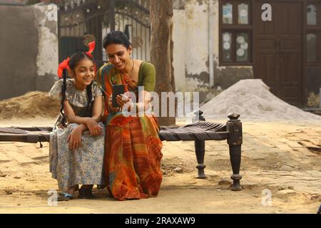 Glückliche indische Landfamilie im Dorf. Mutter und Tochter sitzen zusammen und lächeln auf dem Kinderbett vor ihrem Haus mit dem Handy. Stockfoto