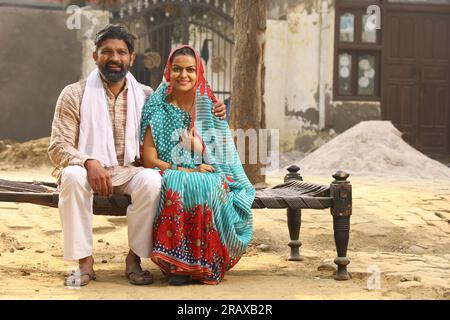 Glückliche indische Landfamilie im Dorf. Mann und Frau sitzen auf einem Kinderbett vor ihrem Vorgarten. Mann in Kurta-Pyjamas und Frau in wunderschöner Saree. Stockfoto