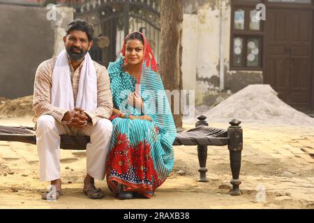 Glückliche indische Landfamilie im Dorf. Mann und Frau sitzen auf einem Kinderbett vor ihrem Vorgarten. Mann in Kurta-Pyjamas und Frau in wunderschöner Saree. Stockfoto