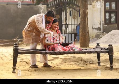 Glückliche indische Landfamilie im Dorf. Mann und Frau sitzen auf einem Kinderbett vor ihrem Vorgarten. Mann in Kurta-Pyjamas und Frau in wunderschöner Saree. Stockfoto