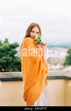 Romantisches Porträt einer schönen Frau auf dem Balkon unter dem Sommerregen, mit einem rothaarigen Strickumhang Stockfoto