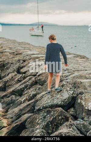 Hübsches kleines Mädchen, das an einem kalten Tag am See spielt, in einem marineblauen Kleid, Rückansicht Stockfoto