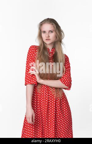Modische junge blonde Frau mit langen Haaren in einem roten Sommerkleid mit Punktmuster auf weißem Hintergrund. 21-jährige Frau, die in die Kamera schaut Stockfoto