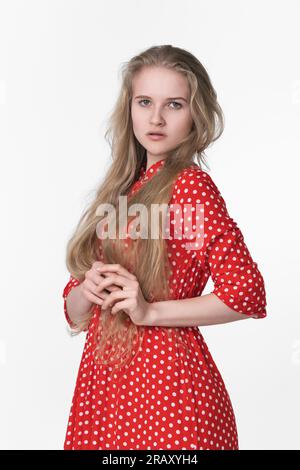 Elegante junge blonde Frau mit langen Haaren in einem roten Sommerkleid mit Punktmuster auf weißem Hintergrund. Weiße ethnische Frau 21 Jahre alt Stockfoto