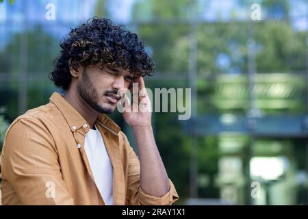 Nahaufnahme. Ein junger Indianer wurde auf der Straße krank. Er hält seinen Kopf mit der Hand, fühlt starke Schmerzen, Druck und Stress. Er schließt seine Augen und Grimaces vor der Spannung. Stockfoto
