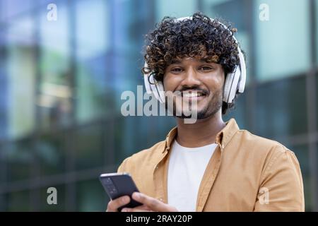 Nahaufnahme. Porträt eines jungen Inders, der Kopfhörer trägt, draußen steht, ein Telefon hält und in die Kamera lächelt. Stockfoto