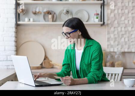 Wunderschöne junge muslimische Frau in Hidschab mit Kopfhörern, die zu Hause in der Küche sitzt und ein Notebook benutzt. Sie hört Musik, ist zufrieden, hebt die Hände hoch, tanzt mit. Stockfoto