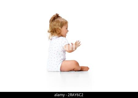 Positives, fröhliches, sorgenfreies kleines Mädchen, das auf dem Boden sitzt und sich vor weißem Studiohintergrund die Hände klatscht Stockfoto