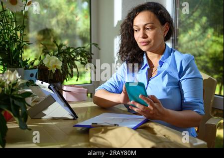 Eine selbstbewusste Geschäftsfrau, Blumendesignerin, die ihr Handy benutzt, arbeitet mit Bestellungen für Blumenarrangements in der Werkstatt, sitzt mit Digi am Schreibtisch Stockfoto