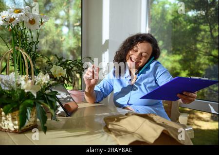 Lächelnde Frau, die mit dem Handy telefoniert, Bestellungen für Blumenarrangements im Blumenladen entgegennimmt, am Schreibtisch in ihrer Werkstatt sitzt, umgeben von Beautifu Stockfoto