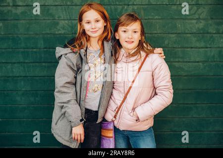Außenporträt von zwei süßen kleinen Mädchen, die warme Jacken tragen, vor grünem Hintergrund posieren, Mode für Kinder Stockfoto